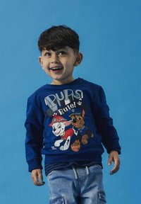 Felpa blu con cani cartone animato, uno in uniforme di polizia, con il testo "I PUPI Regnano!". Abbinata a pantaloni cargo azzurri.