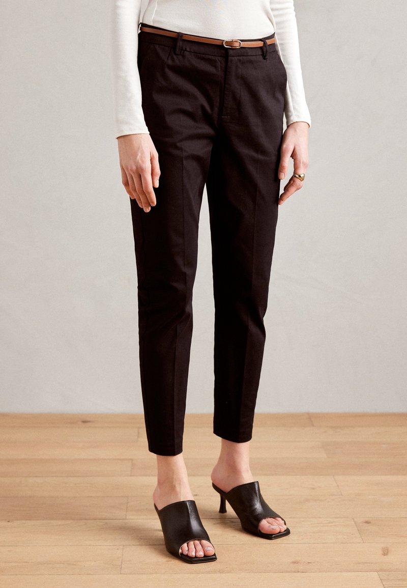 Femme portant un pantalon noir slim raccourci, une ceinture marron, un haut blanc à manches longues, des mules à talons ouvertes noires, debout sur un sol en bois.
