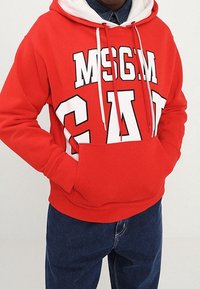 Persona que lleva una sudadera roja de MSGM con cordones blancos y un bolsillo delantero, combinada con jeans de color azul oscuro.