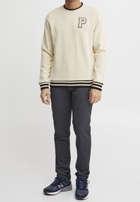 Krémová mikina s černými proužkovanými manžetami a lemem, zdobená vyšívaným písmenem "P." Kombinováno s šedými kalhotami se slim fit střihem a modrou sportovní obuví.