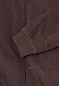 Brown corduroy shirt with a button-down front, horizontal ribbed texture, and fitted cuffs. Includes branded button detail on the front.