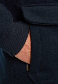 Námořnická vlněná bunda s rukou v boční kapse; má žebrované manžety a nášivku s nápisem "TIME LEGEND" ve zlatém písmu.