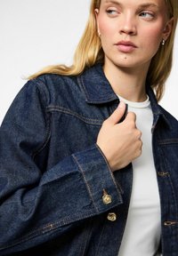 Jeans jakna tamnoplave boje s zlatnim akcentima. Ima šiljastu kragnu, dugmad za zatvaranje i vidljive detalje šivanja.