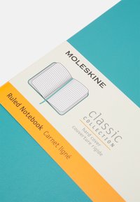 Il quaderno a righe della collezione classica Moleskine presenta una copertina rigida turchese, con un'etichetta arancione che dettaglia le informazioni sul prodotto e un'illustrazione.