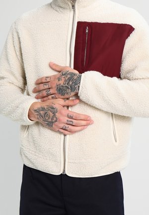 Osoba ubrana w beżową bluzę sherpa z bordową kieszenią na piersi, z założonymi rękami, pokazująca liczne szczegółowe tatuaże na palcach i grzbiecie dłoni.