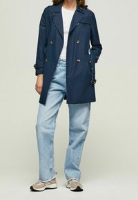 Marinblå trench coat med dubbelknäppning, guldknappar och uppvikta ärmar, kombinerad med ljusblå jeans och flertaliga färgade sneakers.