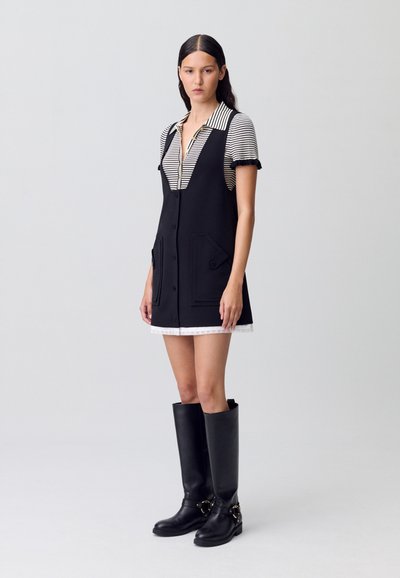 Abito nero senza maniche con tasche frontali e una camicia a righe sottostante. Stivali neri al ginocchio con accenti dorati completano l'outfit.