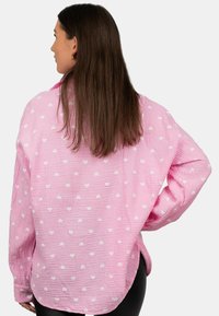 Camicia oversize rosa realizzata in tessuto testurizzato con motivo a cuori bianchi. Presenta maniche lunghe e un orlo arrotondato.