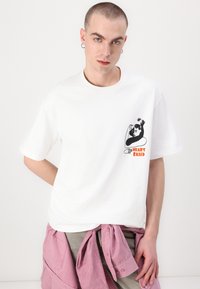 T-shirt bianco in cotone con grafica nera di due volti e la scritta "HEART BASED" in testo arancione sul petto. Maniche corte, vestibilità comoda.