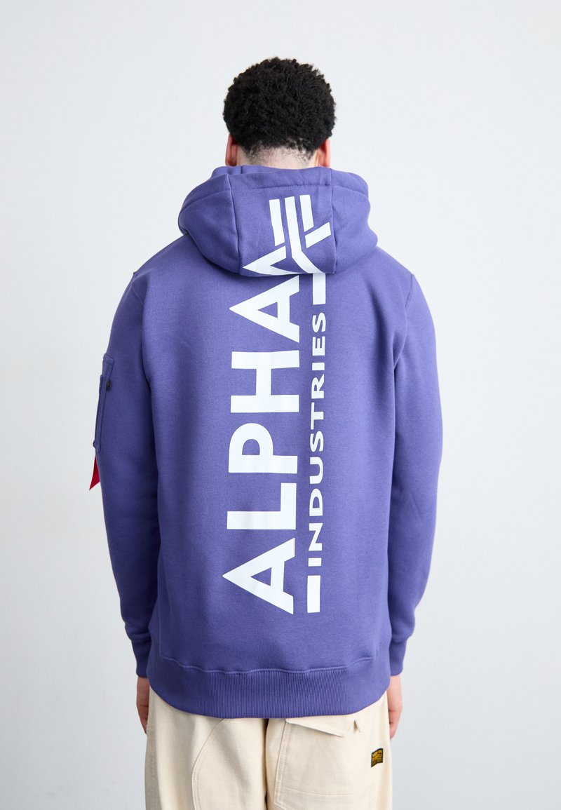 Fialová mikina s kapucňou s bielym nápisom "ALPHA INDUSTRIES" na chrbte, obsahujúca kapucňu a rebrovaný lem. Materiál z bavlneného zloženia, neformálny dizajn.
