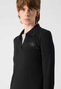 Camisa polo preta de mangas longas com textura canelada, apresentando um colarinho repartido e um pequeno logo cinza da Calvin Klein no lado esquerdo.