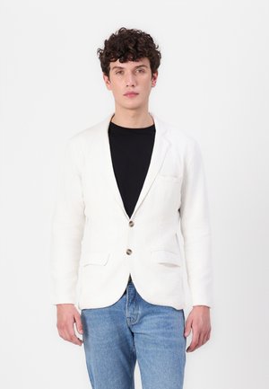 Hvit lin blazer med klaffelapp, to frampocket og to knapper. Båret over en svart skjorte, kombinert med blå jeans.