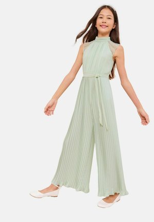 Young girl wearing a sleeveless, pleated light green jumpsuit with a tie waist and white ballet flats, smiling with arms slightly extended.