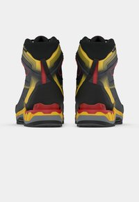 Ténis desportivos de cano alto com um upper em têxtil preto e detalhes em amarelo e vermelho. A sola é grossa, multicolorida, desenhada para proporcionar tração.