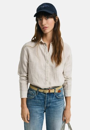 Chemise en lin beige clair avec fermeture à boutons, associée à un jean en denim bleu et une ceinture tissée beige. Porte une casquette marine avec des détails graphiques.