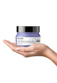 Mascarilla profesional Blondifier de L'Oréal en un tarro morado con tapa negra, sostenido en una mano. El tarro presenta texto blanco que detalla el uso del producto.