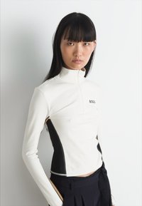 Maglia sportiva a maniche lunghe, bianca, con colletto alto e zip, dotata di pannelli laterali neri e logo "BOSS" sul petto, texture liscia.