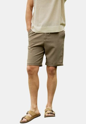 Personne portant un short beige, un haut en maille légère et des sandales marron à enfiler avec des boucles, debout les mains dans les poches.