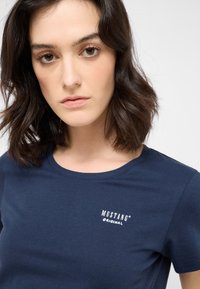 Marineblaue Baumwoll-T-Shirt mit Rundhalsausschnitt und kurzen Ärmeln, das das weiße Logo "MUSTANG ORIGINAL" auf der linken Brustseite zeigt.