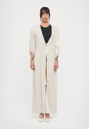 Long, light beige robe with puffed sleeves, open front, and tie closure. Underneath, a fitted black top. Pointed white heels complete the look.