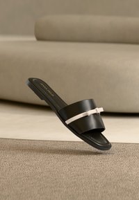 Sandale plate noire avec une large bride et un petit nœud blanc, présentée sur une surface beige texturée avec un fond beige doux.