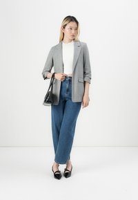 Grijze blazer over een witte top, gecombineerd met high-waisted blauwe jeans en zwarte puntige schoenen. Heeft een kleine zwarte handtas.