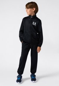 Czarny sportowy dres z zamkiem błyskawicznym i elastycznymi mankietami, z logo na klatce piersiowej; w zestawie z granatowymi trampkami z niebieskimi akcentami.