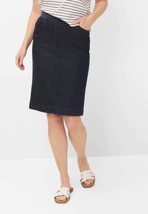 Kvinde iført en mørkeblå knælængde denimskørt, striped skjorte og hvide åbne flade sandaler, står med den ene hånd i lommen.