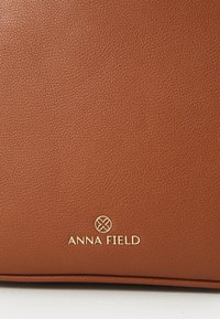 Brun texturerad konstläderhandväska med ett guld "ANNA FIELD" logotypaccent. Har en slät yta och en strukturerad design.