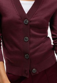 Burgundowy kardigan zapinany na guziki, noszony na białej bluzce, z prążkowanymi brzegami i widocznymi ciemnymi okrągłymi guzikami, noszony przez osobę.