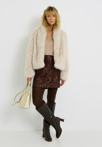 Veste en fausse fourrure beige, haut marron clair, mini-jupe marron foncé pailletée, collants marron, bottes à talons hautes marron jusqu'aux genoux, et sac doré métallisé.