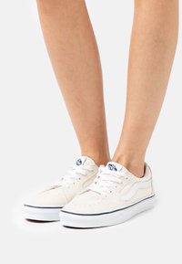 Beige Leinensneaker mit weißen Schnürsenkeln, gepolsterter Zunge und seitlichem Streifen. Ausgestattet mit einem strukturierten Obermaterial und einer kontrastierenden dunklen Sohle.