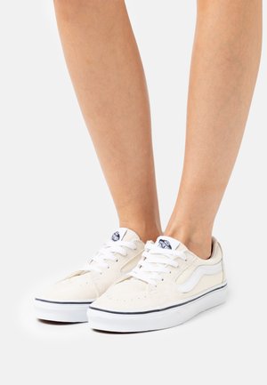 Zapatillas de lona beige con cordones blancos, lengüeta acolchada y raya lateral. Presenta un upper texturizado y suela oscura en contraste.