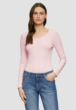 Haut long à manches roses clair avec un décolleté en V, fabriqué en matière douce ; associé à un jeans bleu, se tenant devant un fond gris.