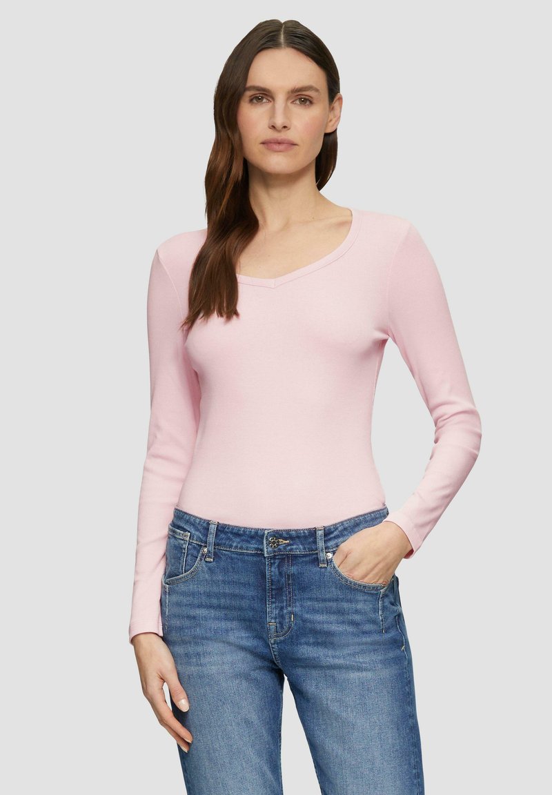 Helles pinkes Langarmshirt mit V-Ausschnitt, aus einem weichen Material; kombiniert mit blauen Jeans, vor einem grauen Hintergrund stehend.