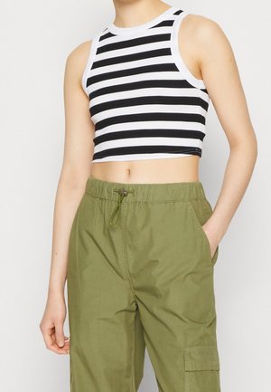 Slanke persoon die een zwart-wit gestreepte cropped tanktop draagt met olijfgroene cargobroek met trekkoord, één hand in de zak.