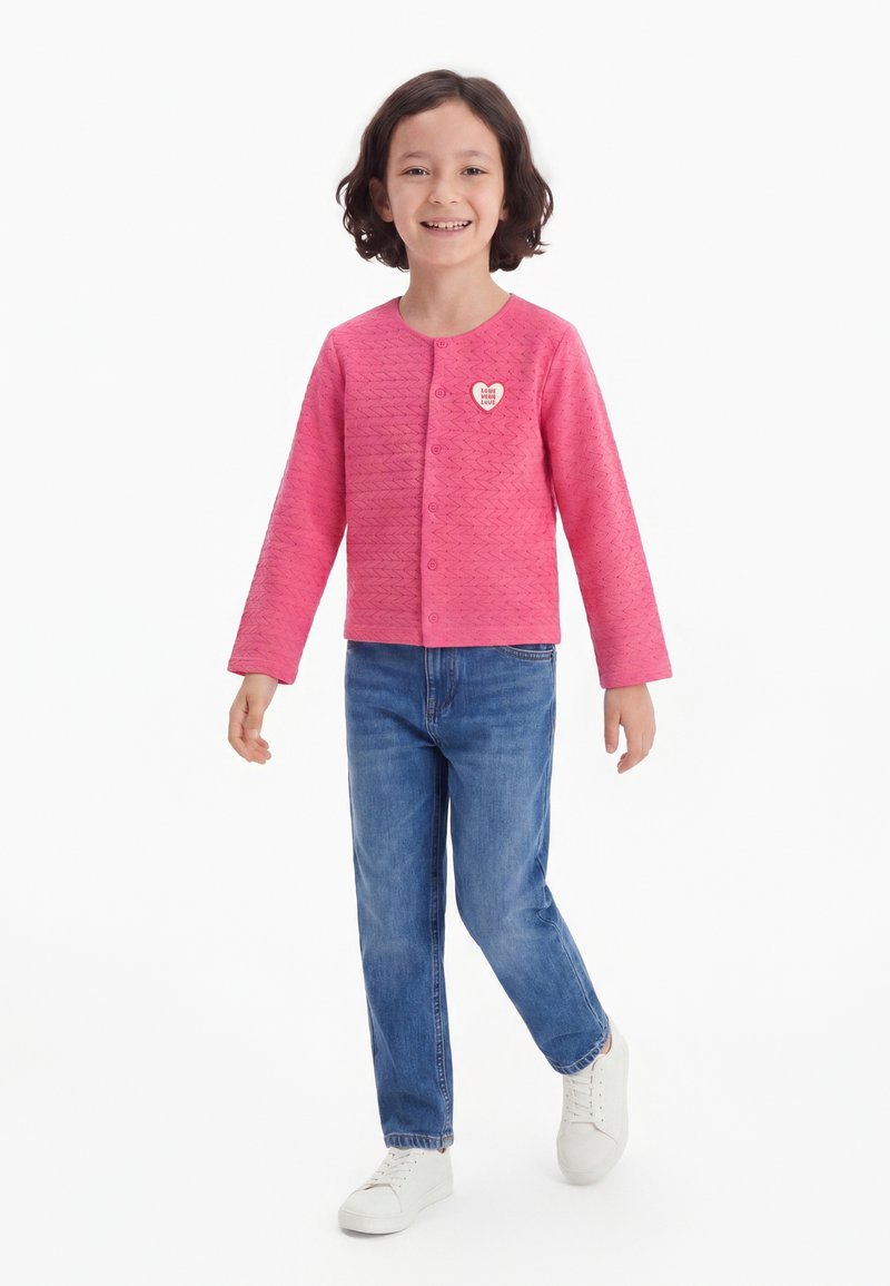 Enfant souriant portant une chemise rose à manches longues boutonnée avec un badge en forme de cœur, un jean bleu et des baskets blanches, debout avec un pied en avant.