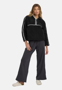 Schwarzer Fleece-Pullover mit einem weißen Reißverschluss und gestreiften Akzenten, kombiniert mit lockeren grauen Hosen und beigen Slippern.