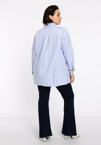 Hellblaue gestreifte Übergröße-Bluse mit hohem Kragen und bestickten Bündchen, kombiniert mit dunkelblauen Schlagjeans und weißen Absätzen.