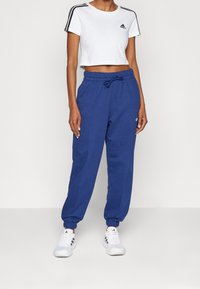 Bílé krátké tričko s černými proužky na rukávech, doplněné modrými tepláky s elastickým pasem a manžetami na kotnících. Bílé tenisky.