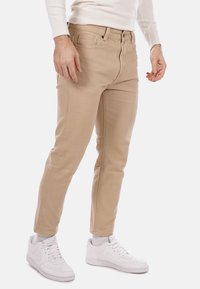 Pantaloni in twill di cotone beige con vestibilità slim, design a cinque tasche e zip frontale. Abbinati a sneakers bianche.