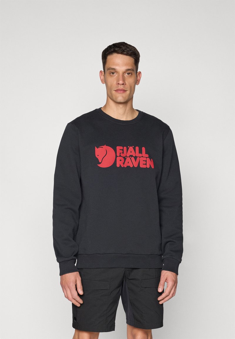 Čierna mikina s červeným logom "FJÄLL RAVEN", okrúhly výstrih, široké manžety a lem. Nosené s čiernymi šortkami. Jednoduché pozadie.