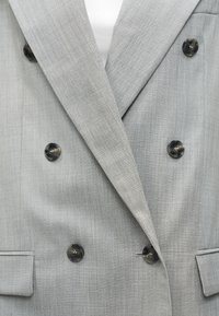 Ανοιχτό γκρι διπλόσαρκο σακάκι με έξι μαύρα κουμπιά και δύο τσέπες με καπάκι, φορεμένο πάνω από λευκό τοπ.