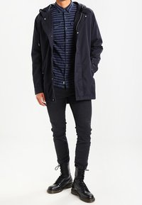 Giacca impermeabile navy con cappuccio, zip frontale e bottoni a scatto sopra una camicia a righe con bottoni. Jeans neri aderenti e stivali con lacci.