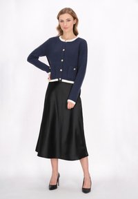 Cardigan a maglia blu navy con bordi color crema, dotato di bottoni dorati e due tasche frontali; indossato su una gonna di raso nera e tacchi.