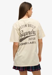Camiseta de algodón color crema con un gran estampado gráfico negro en la parte posterior, con el texto "Superdry Japan Copper Label". Mangas cortas.