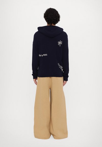 Sweatshirt à capuche bleu marine avec un design de palmier brodé et des détails textuels ; associé à un pantalon beige à jambes larges en tissu doux.