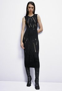 Abito smanicato in maglia nera con motivi verticali e tagli, che arriva fino a metà polpaccio, abbinato a stivali neri aderenti al ginocchio.