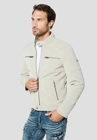 Giacca in finto camoscio grigio chiaro con colletto alto, tasche con cerniera e dettagli a coste sulle spalle, abbinata a jeans blu strappati.