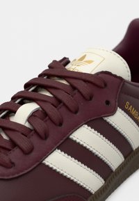 Nærbillede af bordeaux Adidas Samba sneakers med cremefarvede striber, bordeaux snørebånd og guld logo på tungen og siden.
